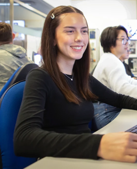 A student smiling while working at a computer in a CAD classroom, surrounded by classmates focused on their designs. A student smiling while working at a computer in a CAD classroom, surrounded by classmates focused on their designs.